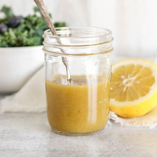 Maple Dijon dressing and a spoon in mason jar with a bowl of greens and a sliced lemon on the background.