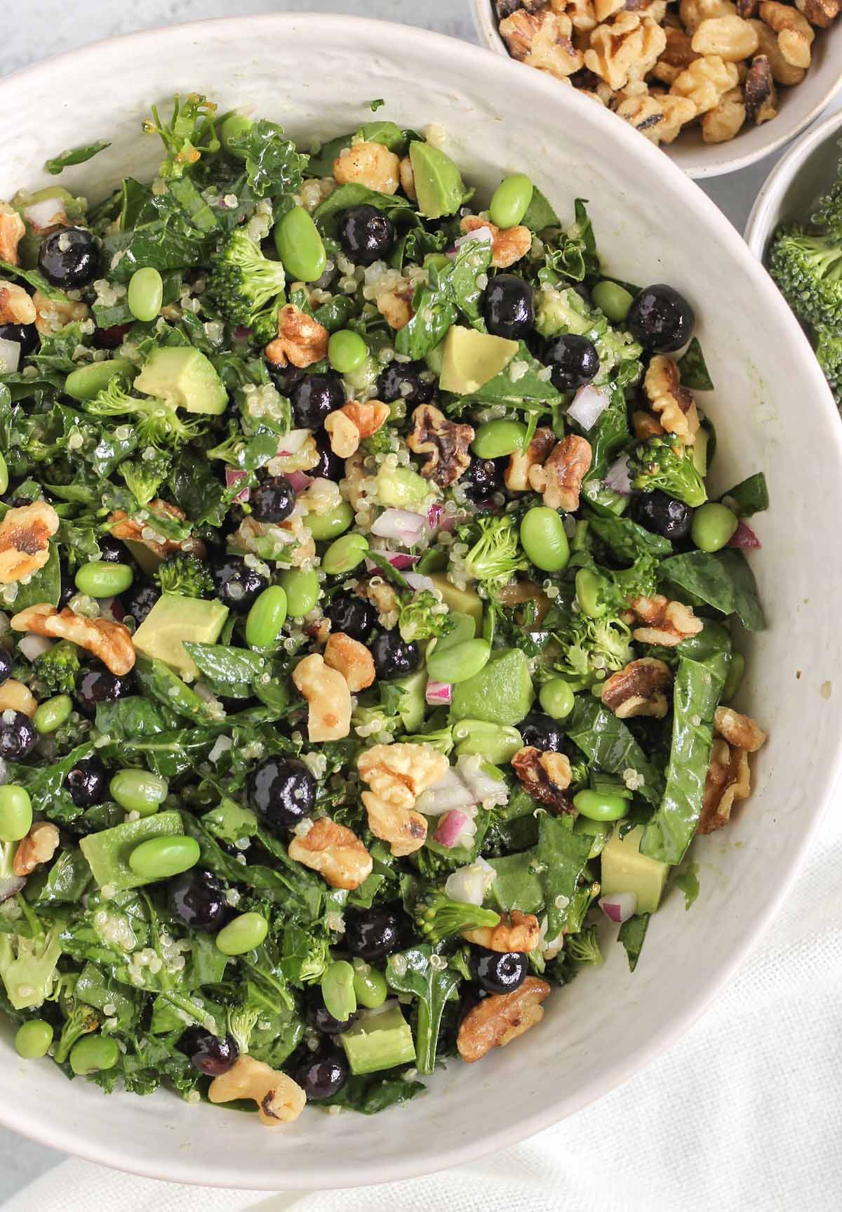 Superfood salad in a large white serving bowl with a bowl of walnuts and broccoli on the side.