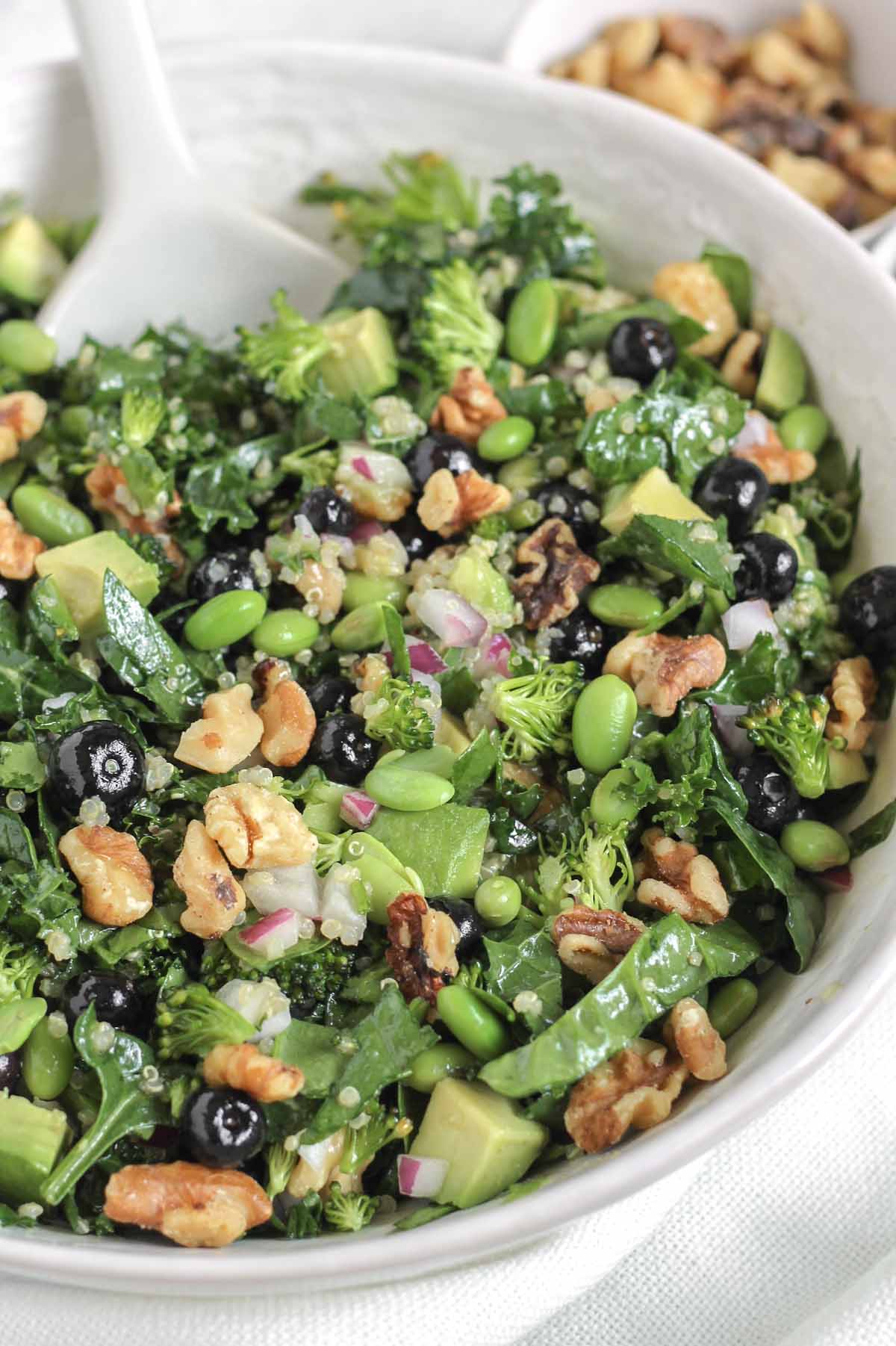 Superfood salad in a large white bowl with a white serving utensils and a bowl of walnuts in the background.