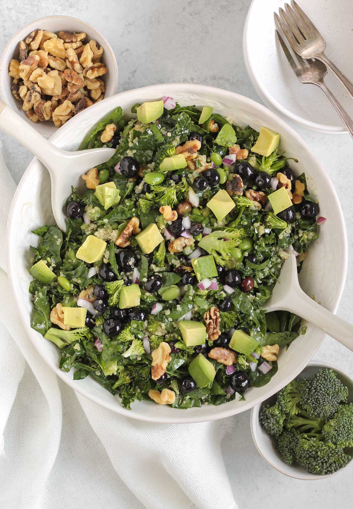 Superfood salad with blueberries, broccoli, walnuts, and avocado in a white serving bowl with white salad utensils with bowls of broccoli and walnuts off to the side.