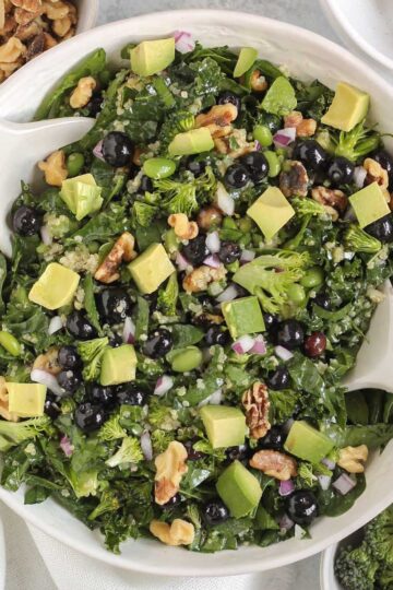 Superfood salad with blueberries, broccoli, walnuts, and avocado in a white serving bowl with white salad utensils.