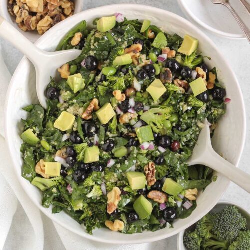 Superfood salad with blueberries, broccoli, walnuts, and avocado in a white serving bowl with white salad utensils.