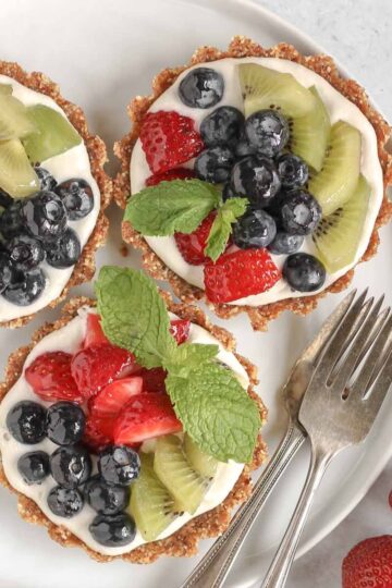 Three mini fruit tarts on a small white plate with 2 vintage forks on the plate and fresh berries off to the side.