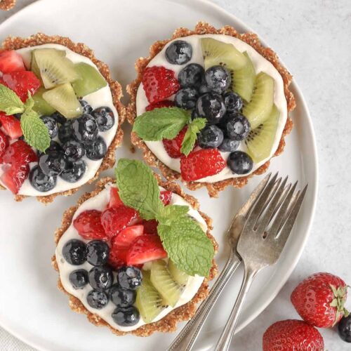 Three mini fruit tarts on a small white plate with 2 vintage forks on the plate and fresh berries off to the side.