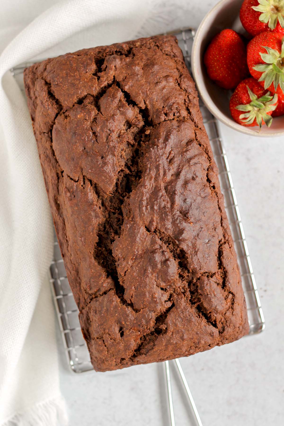 Unsliced loaf of chocolate protein bread on a rectangular cooling rack with a bowl of strawberries on the side.