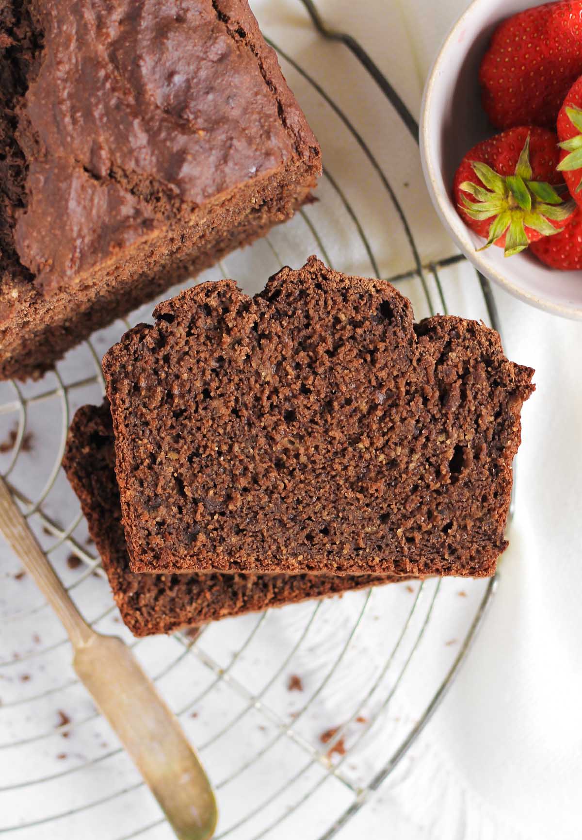 Two slices stacked of chocolate protein banana bread on a round wire cooling rack with the full loaf and a bowl of strawberries to the side.