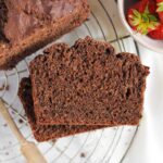 Two slices stacked of chocolate protein banana bread on a round wire cooling rack with a bowl of strawberries to the side.