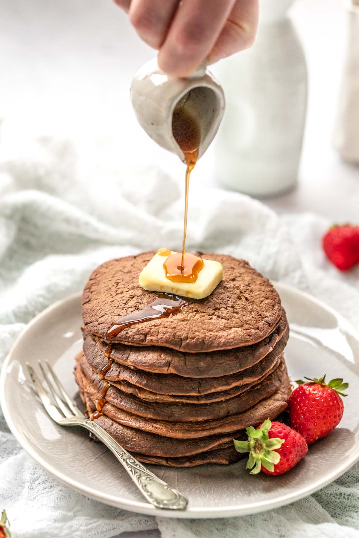 Syrup being poured over a stack of chocolate pancakes topped with a butter square and strawberries on the side.