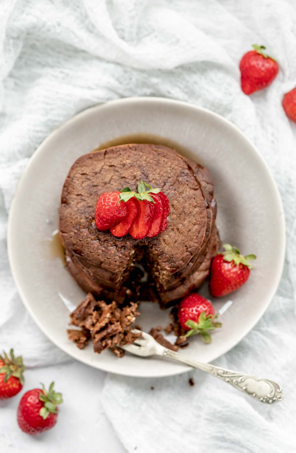 Overhead view of chocolate protein pancakes and strawberries on a plate with a small triangle cut from the pancakes.