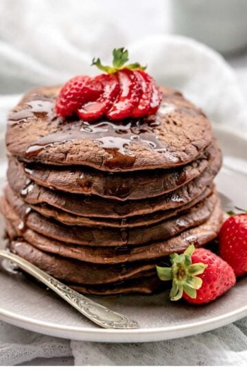 Chocolate pancakes topped with strawberries and dripping with syrup on a plate with a vintage fork.