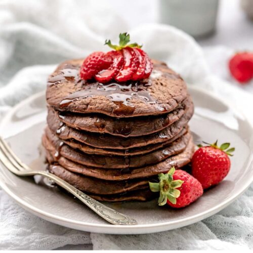 Chocolate pancakes topped with strawberries and dripping with syrup on a plate with a vintage fork.