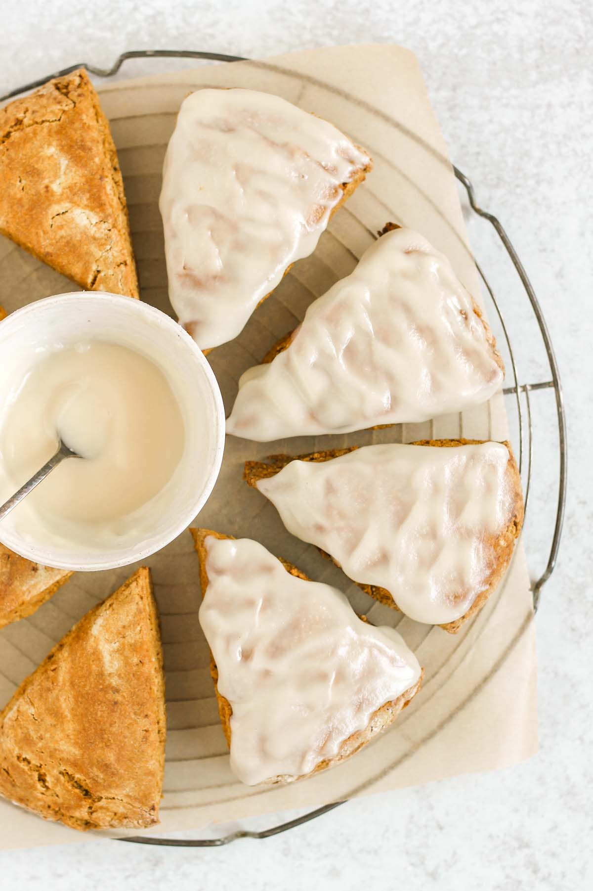 Partially frosted pumpkin scones with a bowl of icing on a round cooling rack.