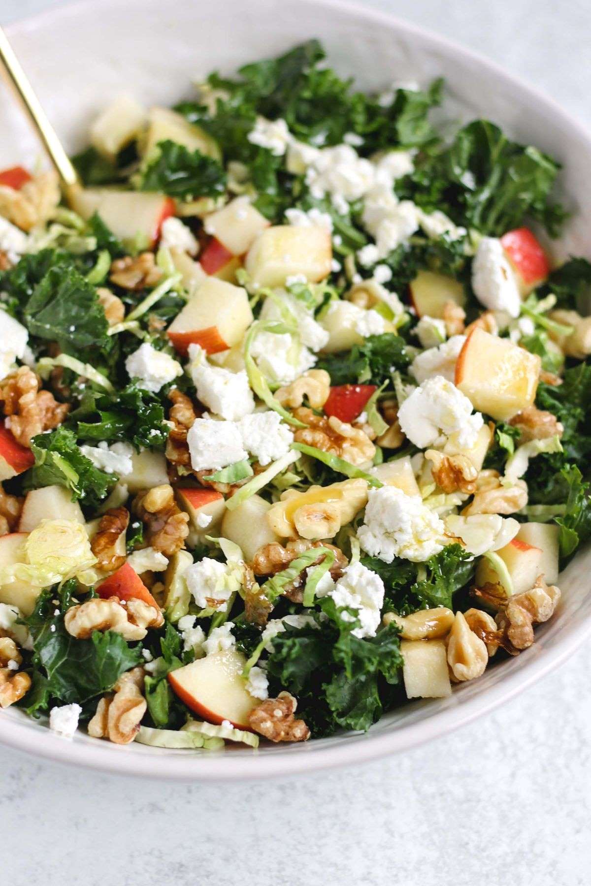 Kale salad with shaved Brussels sprouts topped with apples, walnuts, and goat cheese crumbles in a white serving bowl.