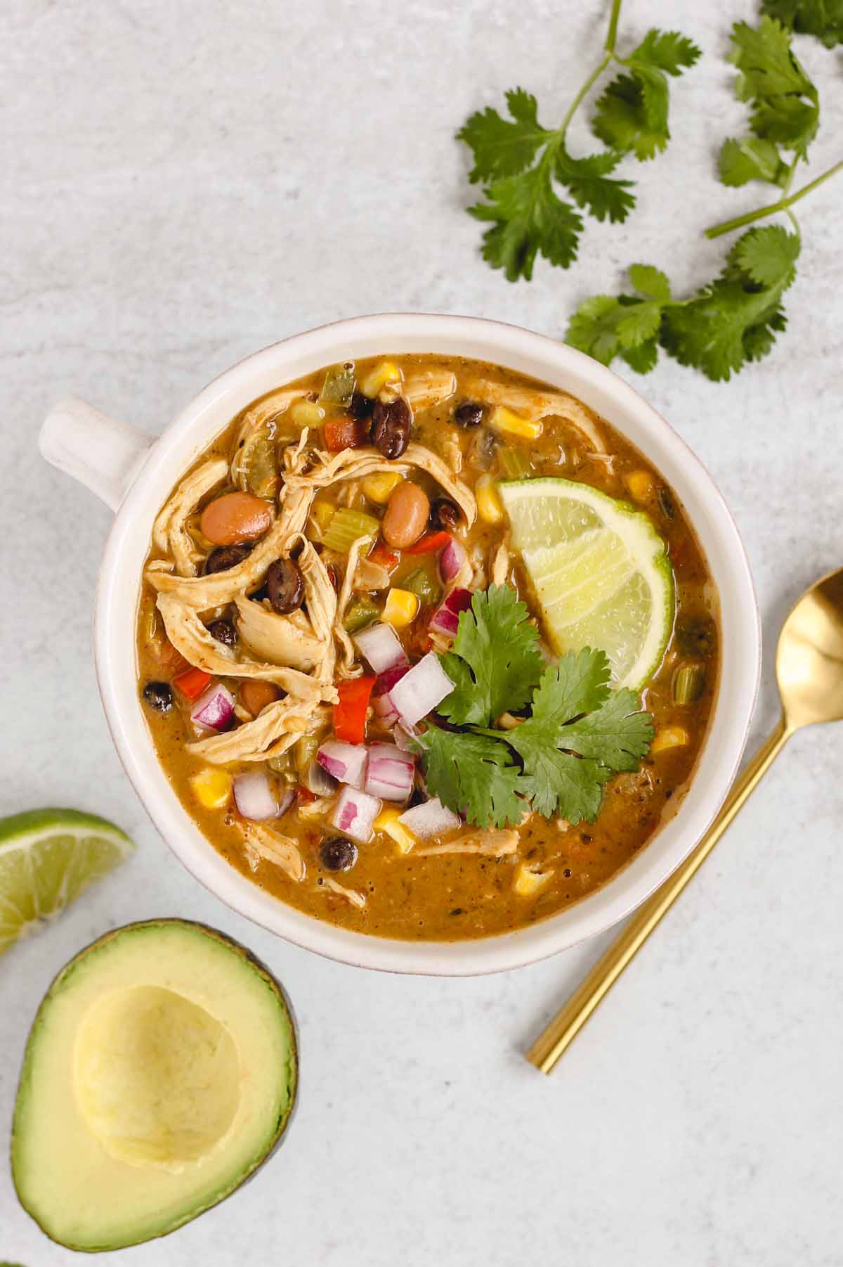 Bowl of chicken chili topped with fresh cilantro and a lime wedge with a gold spoon and avocado half on the side.