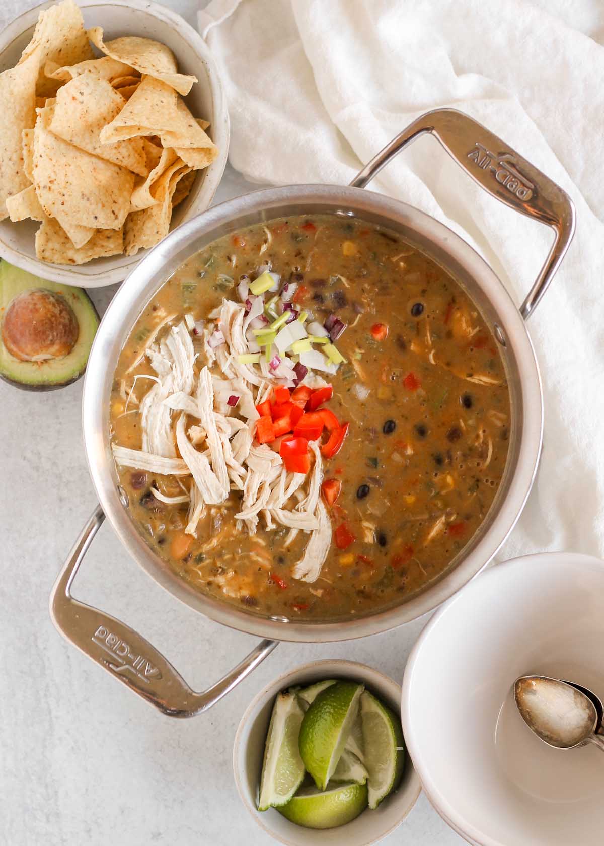 Stainless steel stock pot full of chicken chili with bowls, lime wedges, avocado, and chips on the side of the pot.