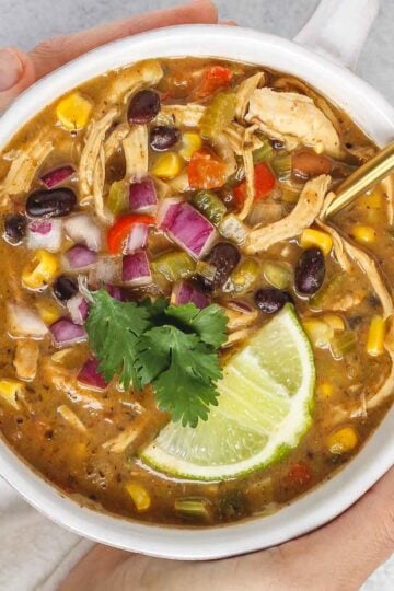 Hands holding a bowl of chicken chili topped with fresh cilantro and a lime wedge.