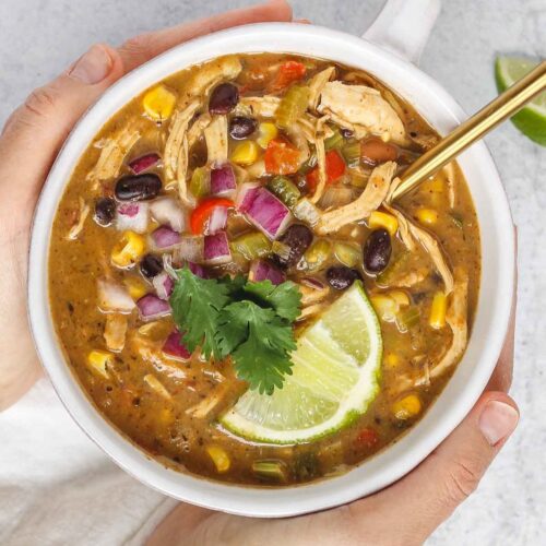 Hands holding a bowl of chicken chili topped with fresh cilantro and a lime wedge.