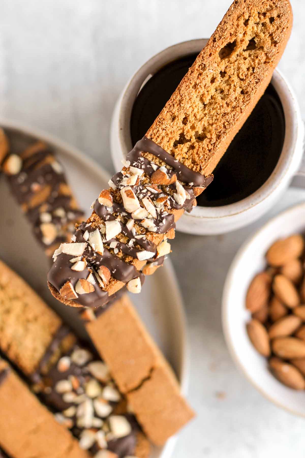 Single biscotti drizzled with chocolate and almond pieces laying onto of a cup of coffee with additional biscotti in the background.