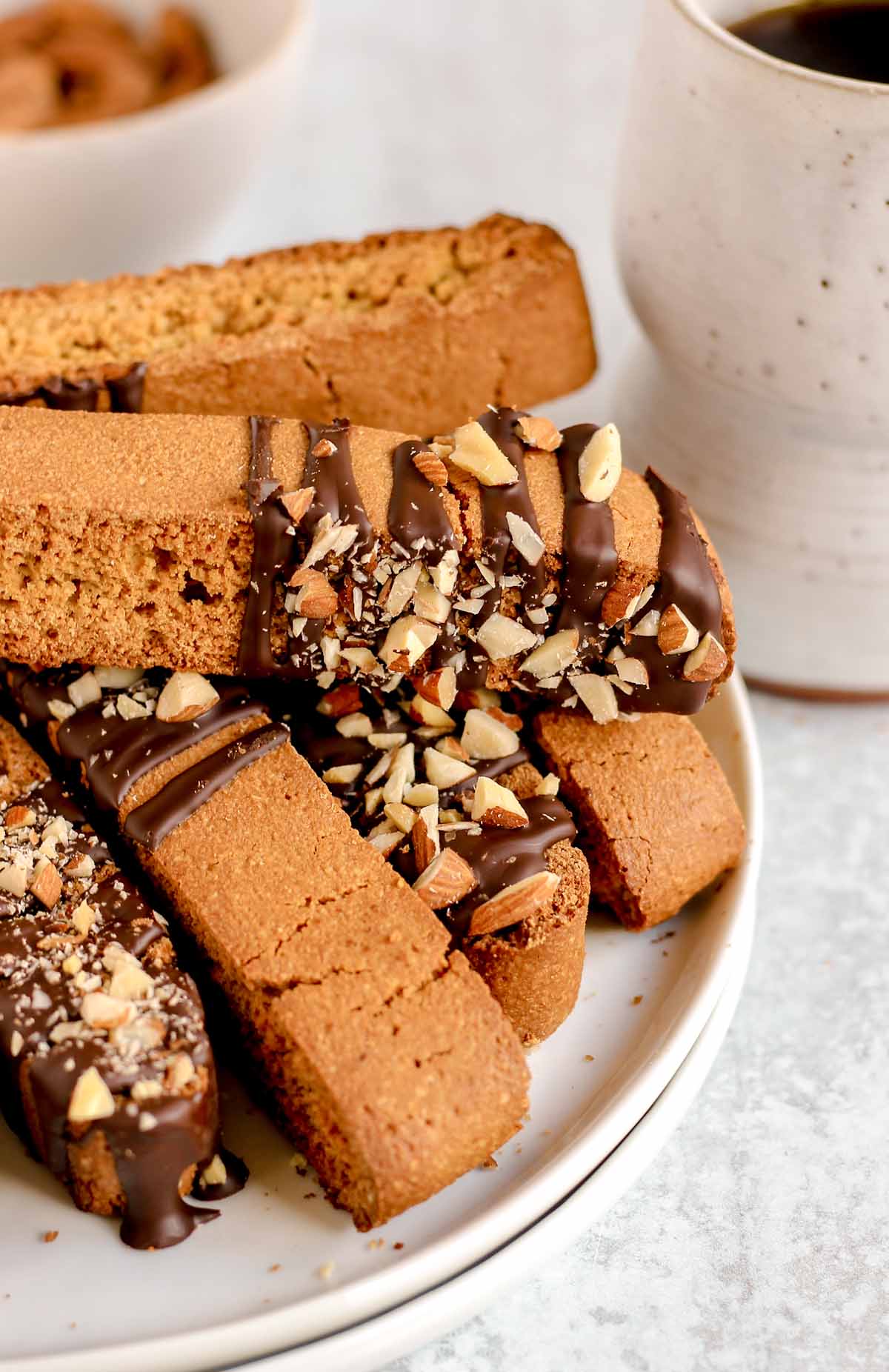 Biscotti drizzled with chocolate and almond pieces stacked on a plate with a cup of coffee in the background.
