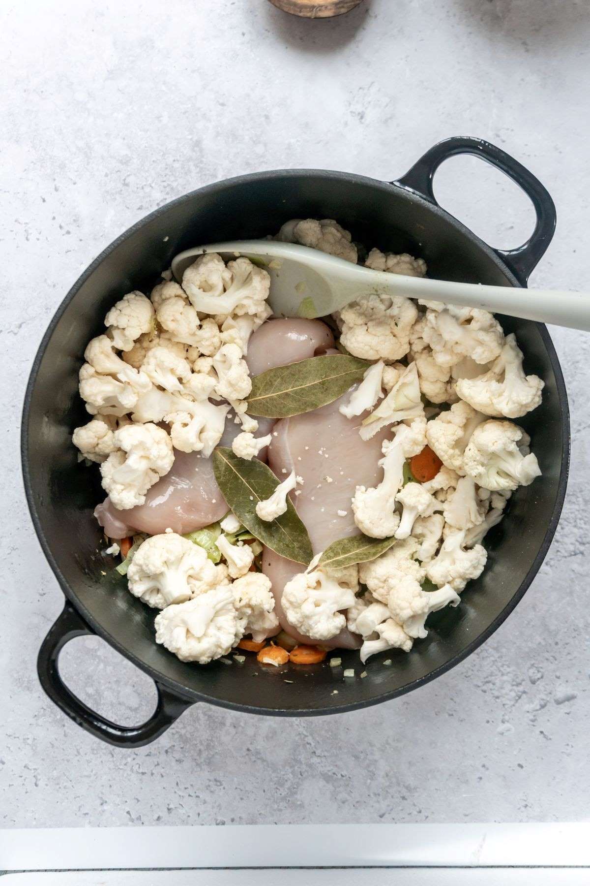 Veggies, raw chicken breasts, cauliflower florets, and bay leaves in a large black stock pot.