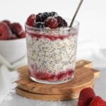 Glass cup, on a small round wooden cutting board, filled with overnight oats topped with thawed frozen fruit with a vintage spoon in the glass.
