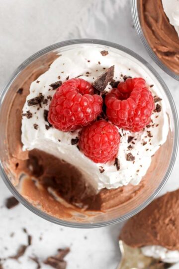 Chocolate mousse topped with whipped cream and raspberries in a small serving dish with a spoonful taken from the dish.