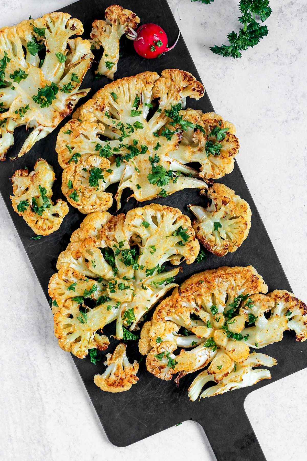 Roasted cauliflower steaks sprinkled with fresh parsley on a black cutting board.