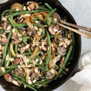 Green beans with mushrooms and onions topped with sliced almonds in a cast iron skillet with a vintage serving spoon and fork in the skillet.