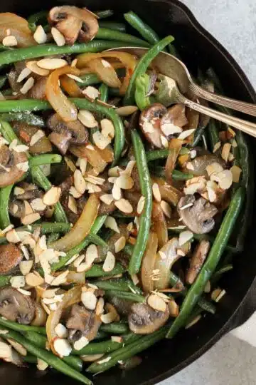 Green beans with mushrooms and onions topped with sliced almonds in a cast iron skillet with a vintage serving spoon and fork in the skillet.