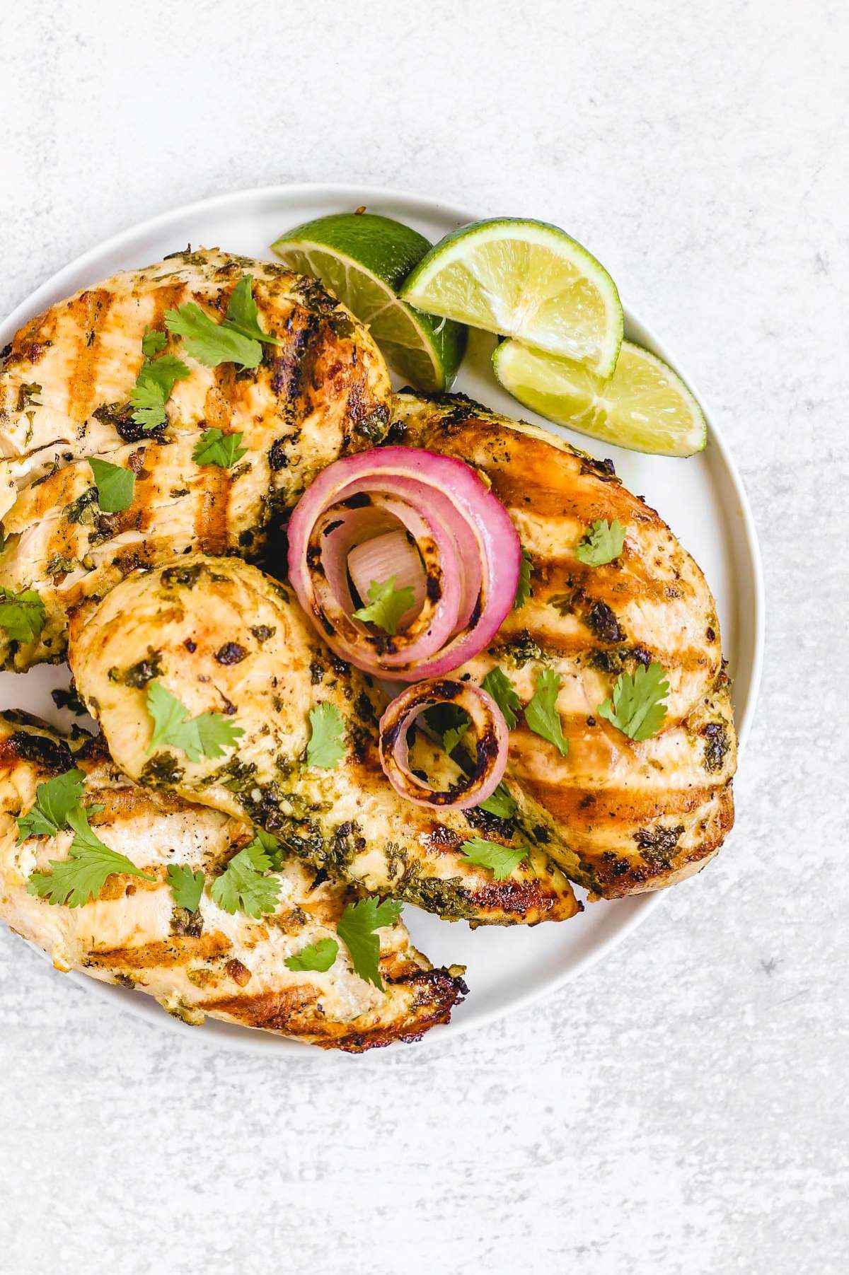 Four grilled cilantro lime chicken breasts with grilled onions and lime slices on a round plate.