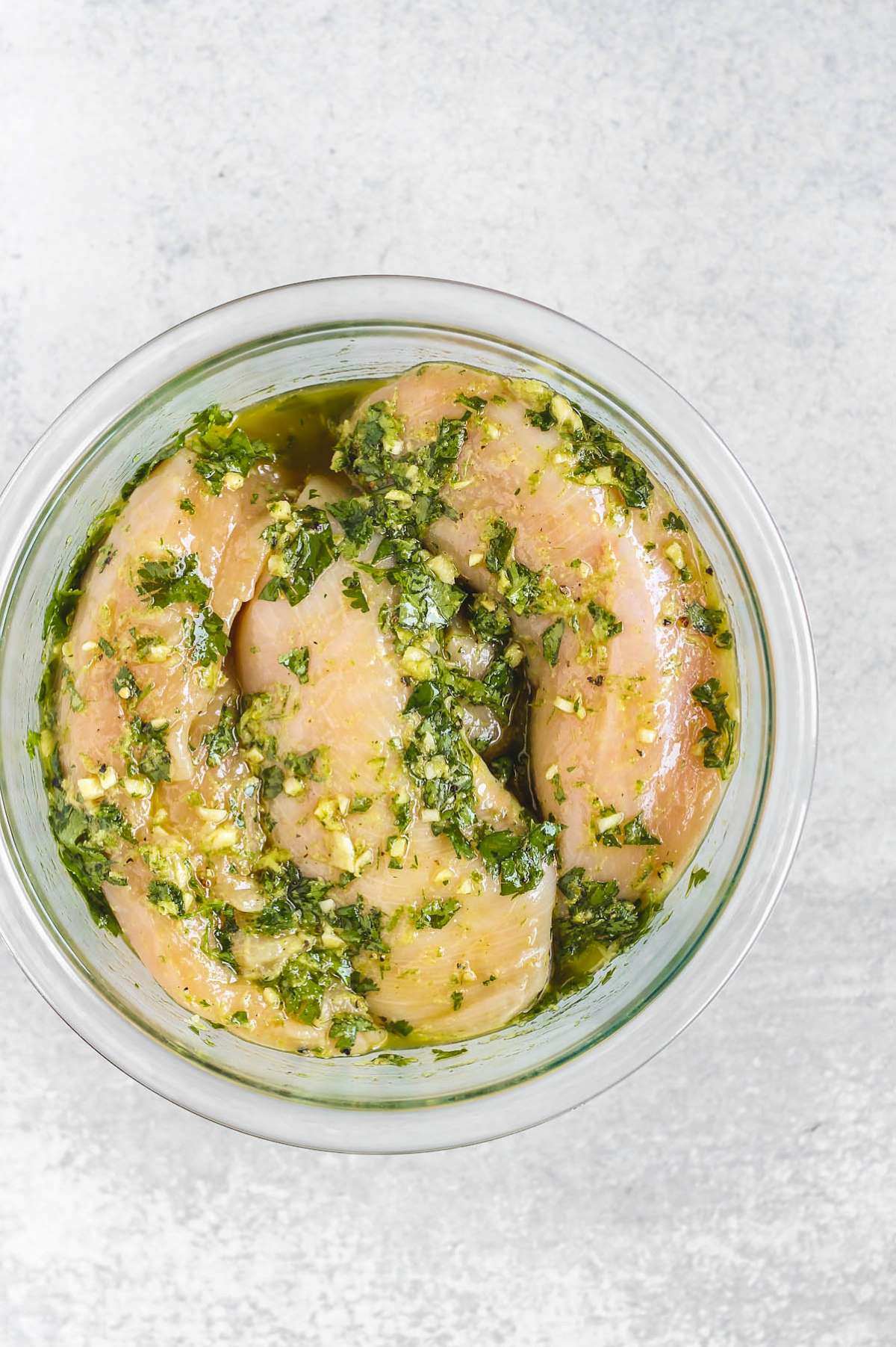 Raw chicken breasts sitting in a cilantro lime marinade in a glass bowl.