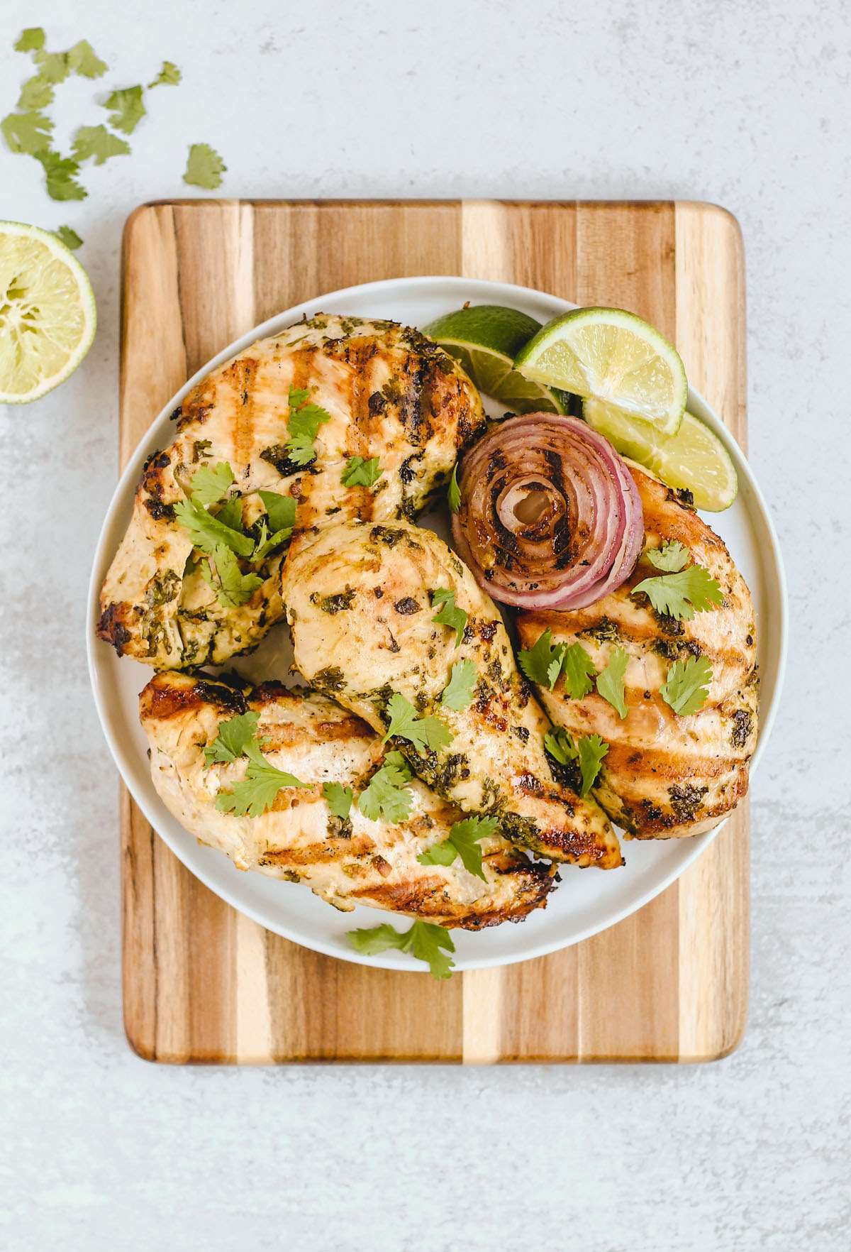 Four grilled cilantro lime chicken breasts with grilled onions and lime slices on a round plate on top of a wood cutting board.