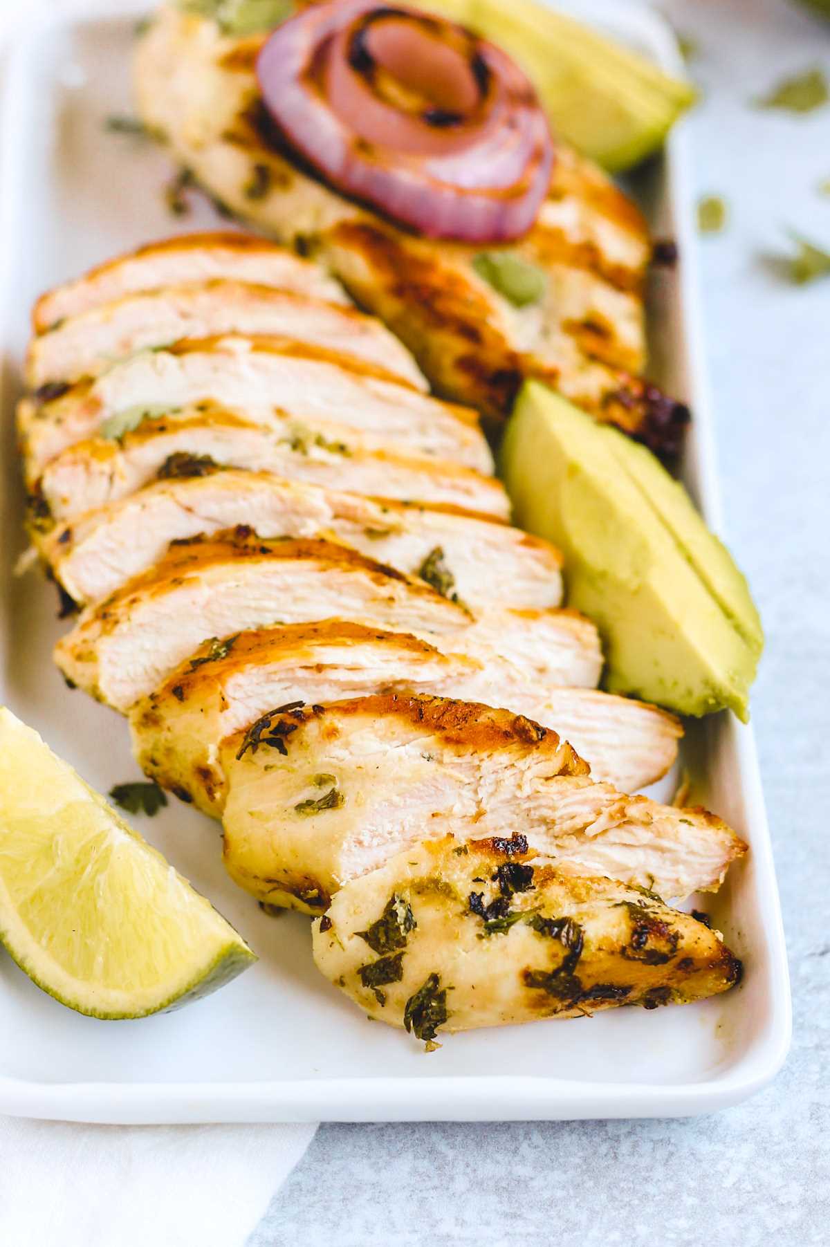 Cooked, and sliced, cilantro lime chicken breast on a rectangular plate with lime wedge and sliced avocado on the side.