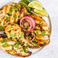 Four grilled cilantro lime chicken breasts with grilled onions and lime slices on a round plate.