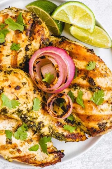 Four grilled cilantro lime chicken breasts with grilled onions and lime slices on a round plate.
