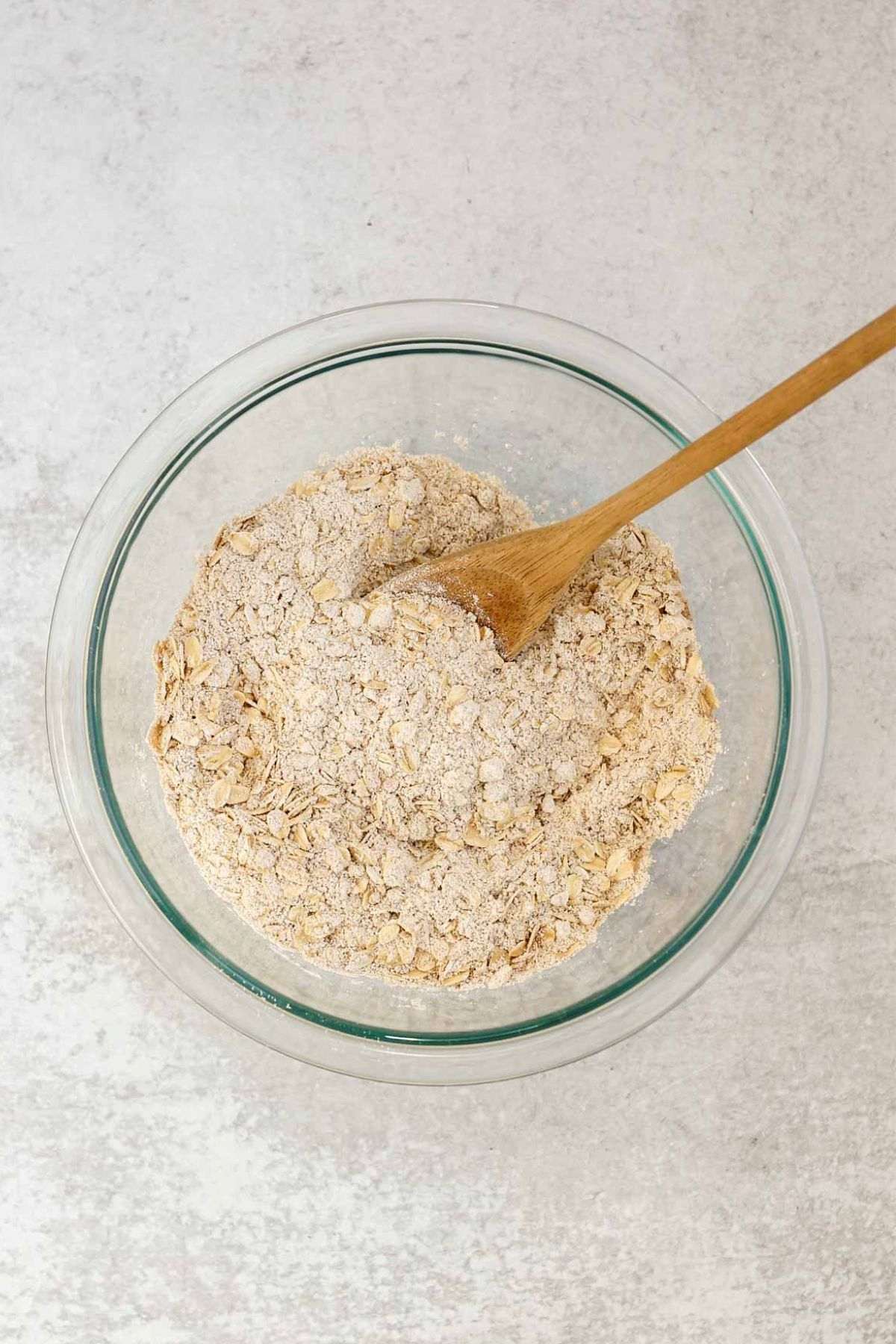 Dry ingredients needed for carrot cake oatmeal cookies in a glass mixing bowl with a wooden spoon.