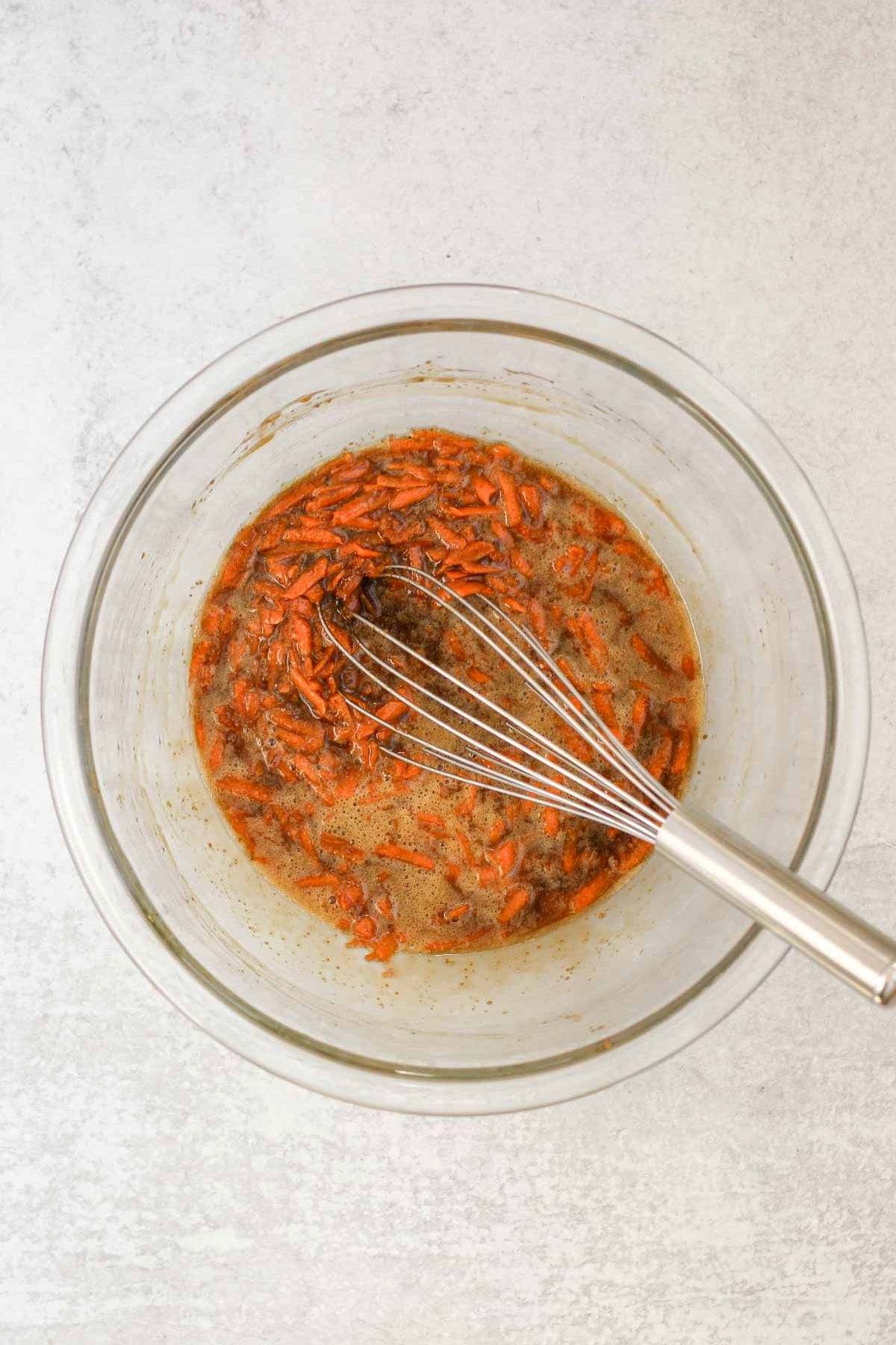 Wet ingredients, plus shredded carrots, needed for oatmeal carrot cake cookies in a glass mixing bowl with a wire whisk.