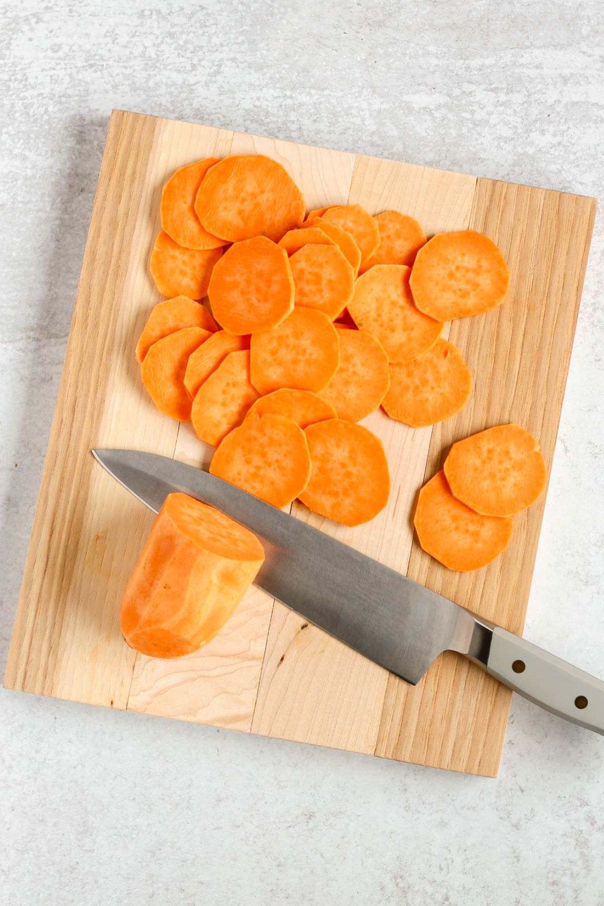 Sliced sweet potatoes with a large knife on a wood cutting board.