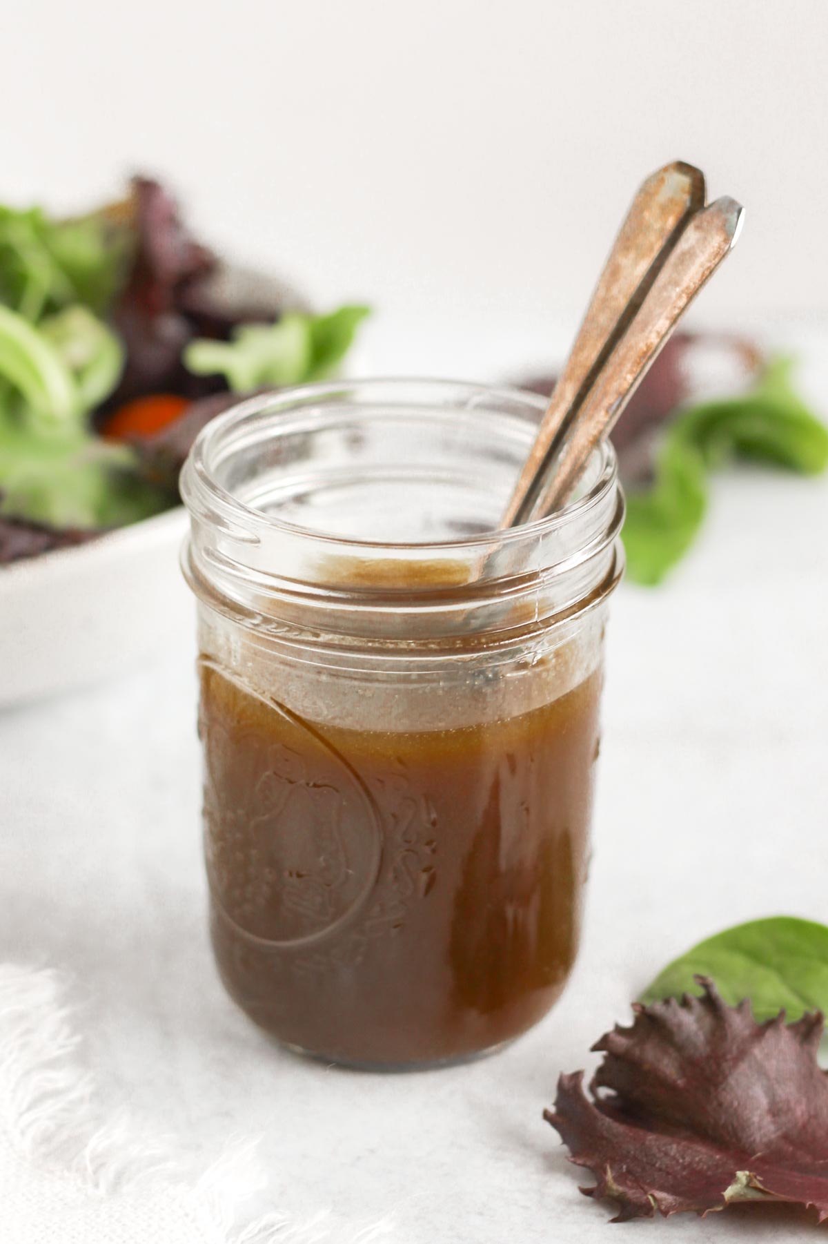 Mason jar with two spoons filled with balsamic vinaigrette with a bowl of mixed greens in the background.