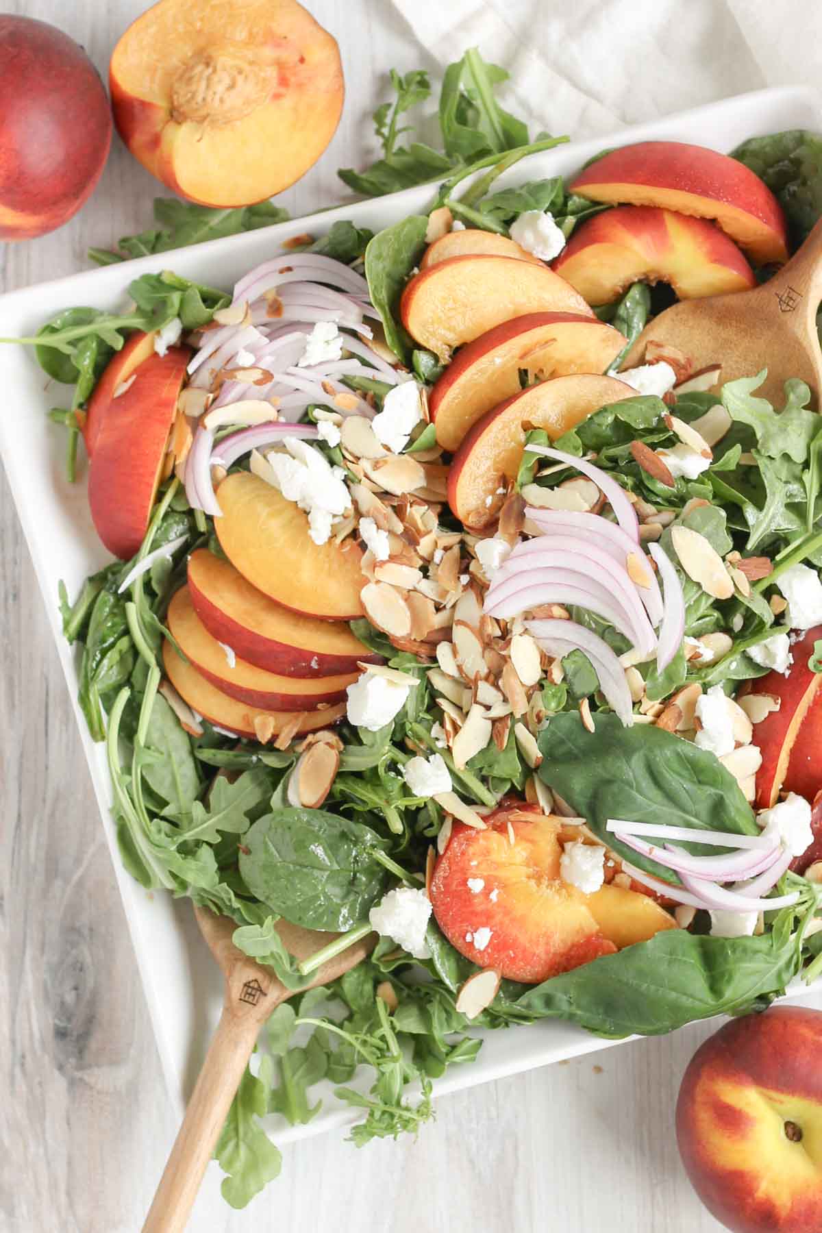 Wooden salad tongs mixing together a peach and goat cheese salad on a square plate with peaches on the sides.