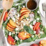 Peach and goat cheese salad with toasted almonds and sliced onions on a square plate with a side of balsamic vinaigrette.