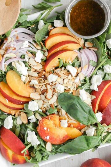 Peach and goat cheese salad with toasted almonds and sliced onions on a square plate with a side of balsamic vinaigrette.
