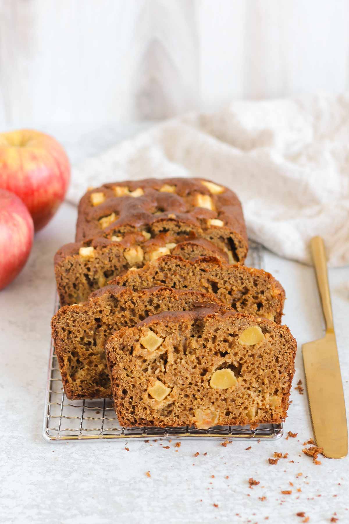 Slices of healthy apple bread on a rectangular wire cooling rack with a gold knife on the side and apples in the background.