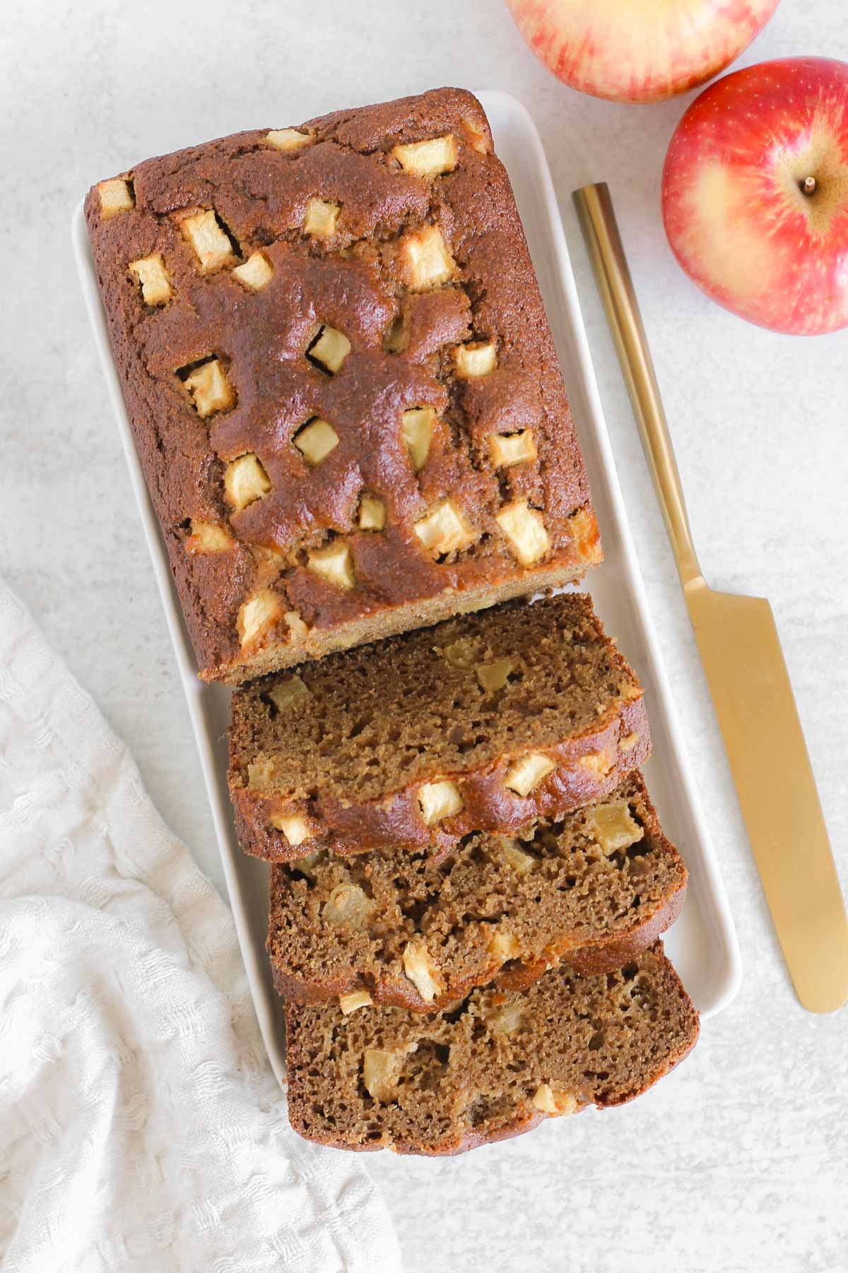 Half sliced loaf of apple bread on a rectangular plate with a gold knife and apples on the side.