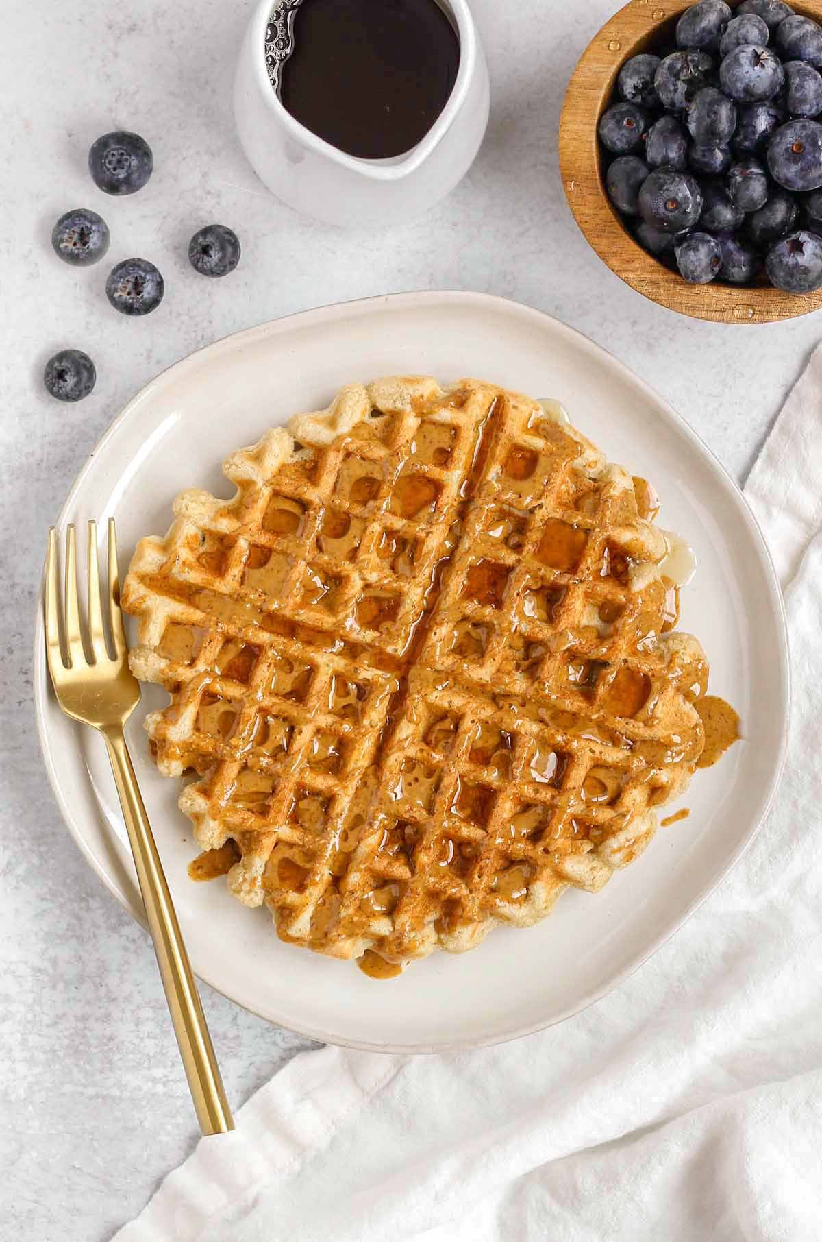 Single waffle on a plate with almond butter and syrup drizzled over the top with blueberries and syrup off to the side.