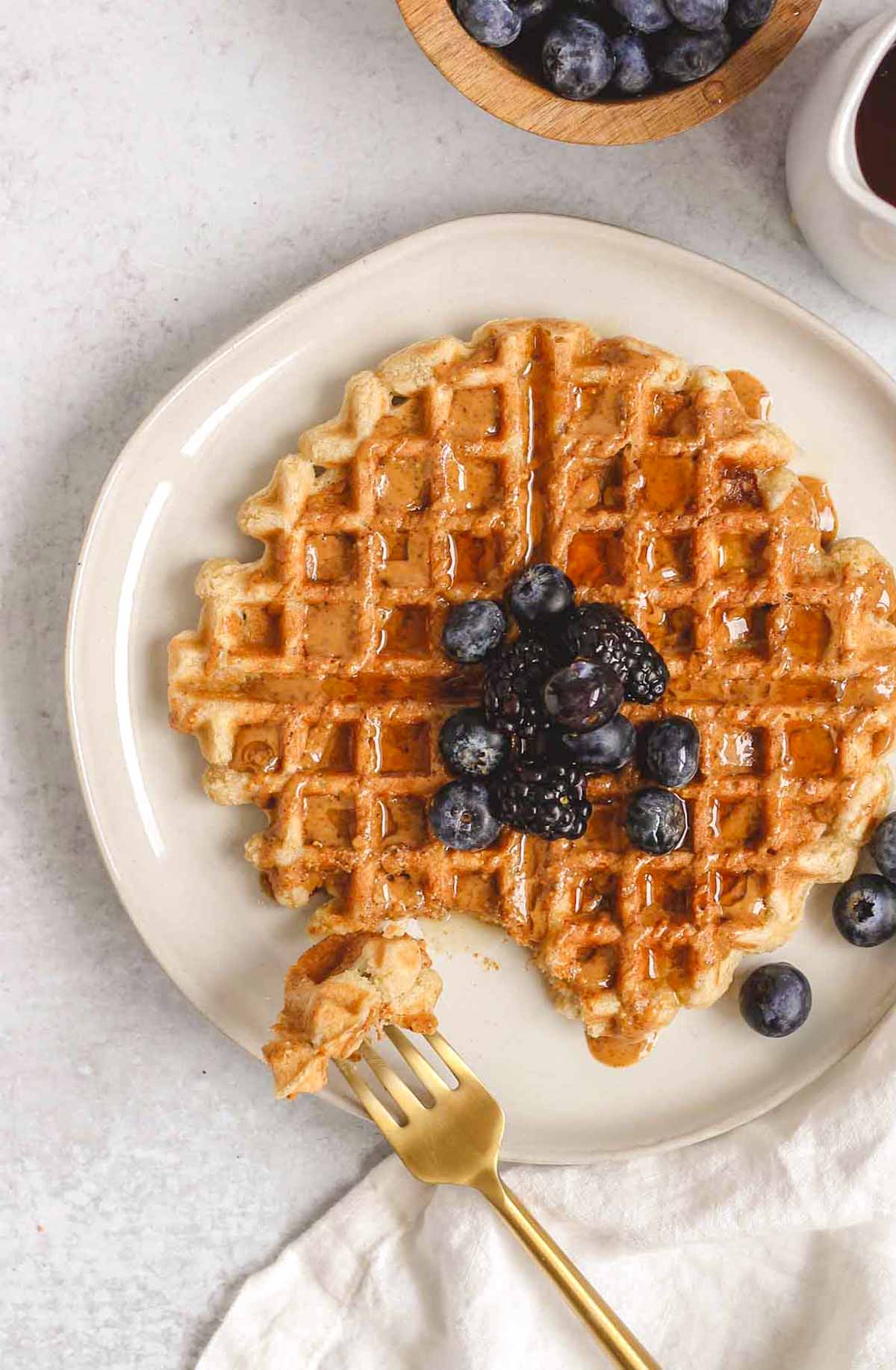 Single waffle topped with berries and drizzled with almond butter and syrup with a gold fork taking a bite from the waffle.