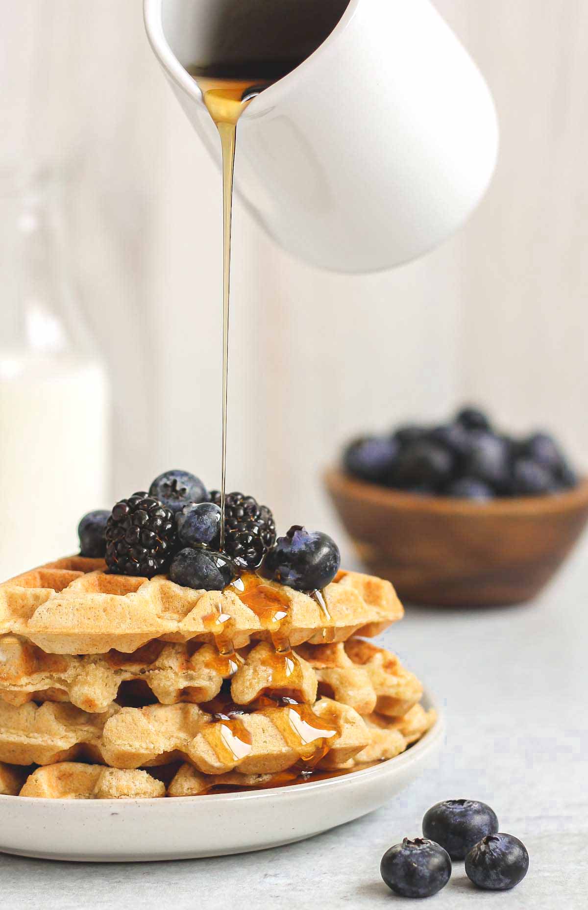 Syrup being poured over a stack of waffles topped with fresh blackberries and blueberries.