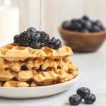 Syrup being poured over a stack of waffles topped with fresh blackberries and blueberries.