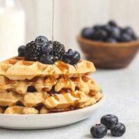 Syrup being poured over a stack of waffles topped with fresh blackberries and blueberries.