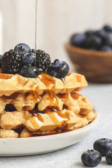Syrup being poured over a stack of waffles topped with fresh blackberries and blueberries.